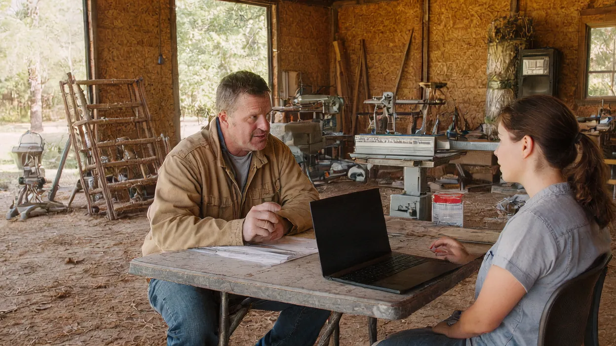 A contractor and a web consultant talk over a laptop at a table in his woodshop.