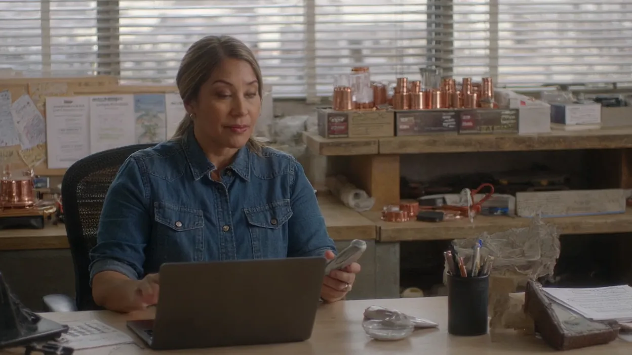 An office manager looks at her phone beside a laptop at her desk, waiting for a test form to come through.
