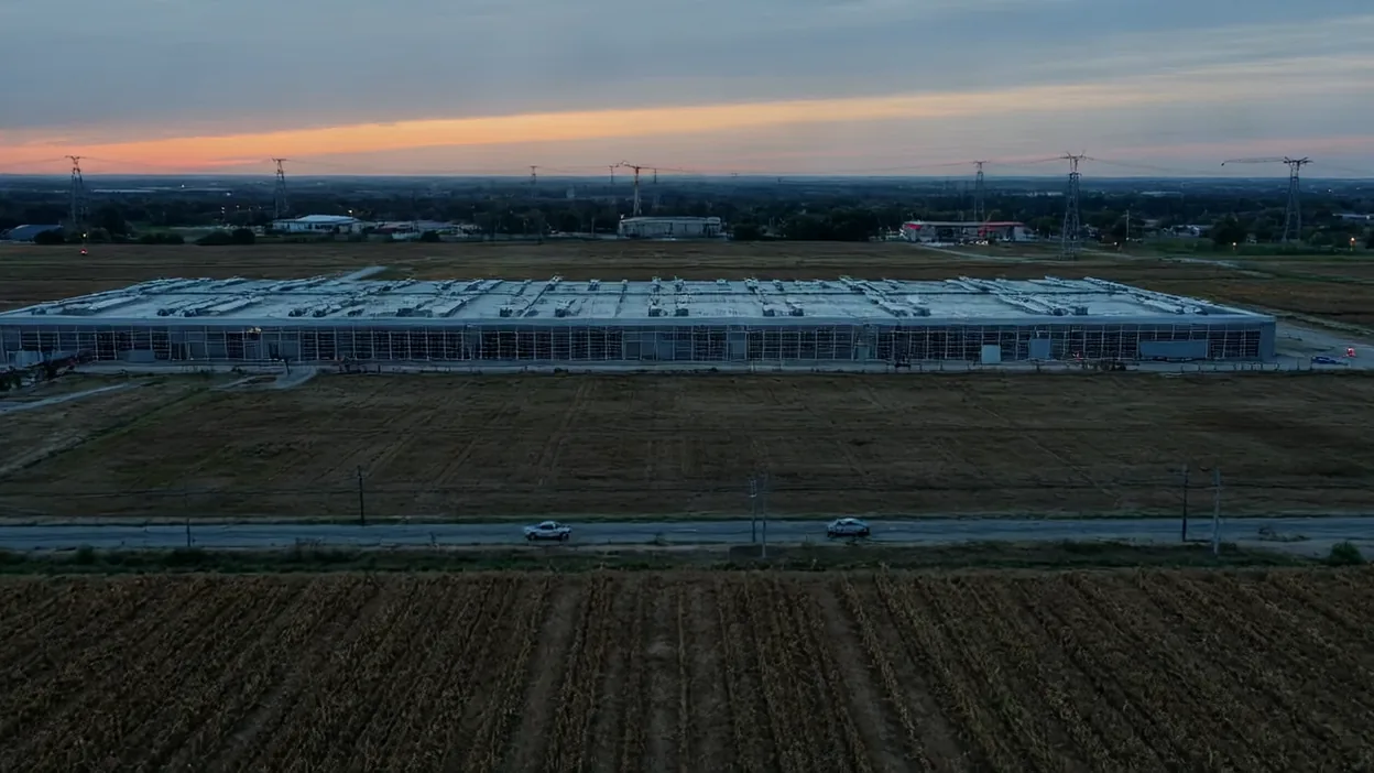 A large, low data center building surrounded by farm fields and power lines at dusk.