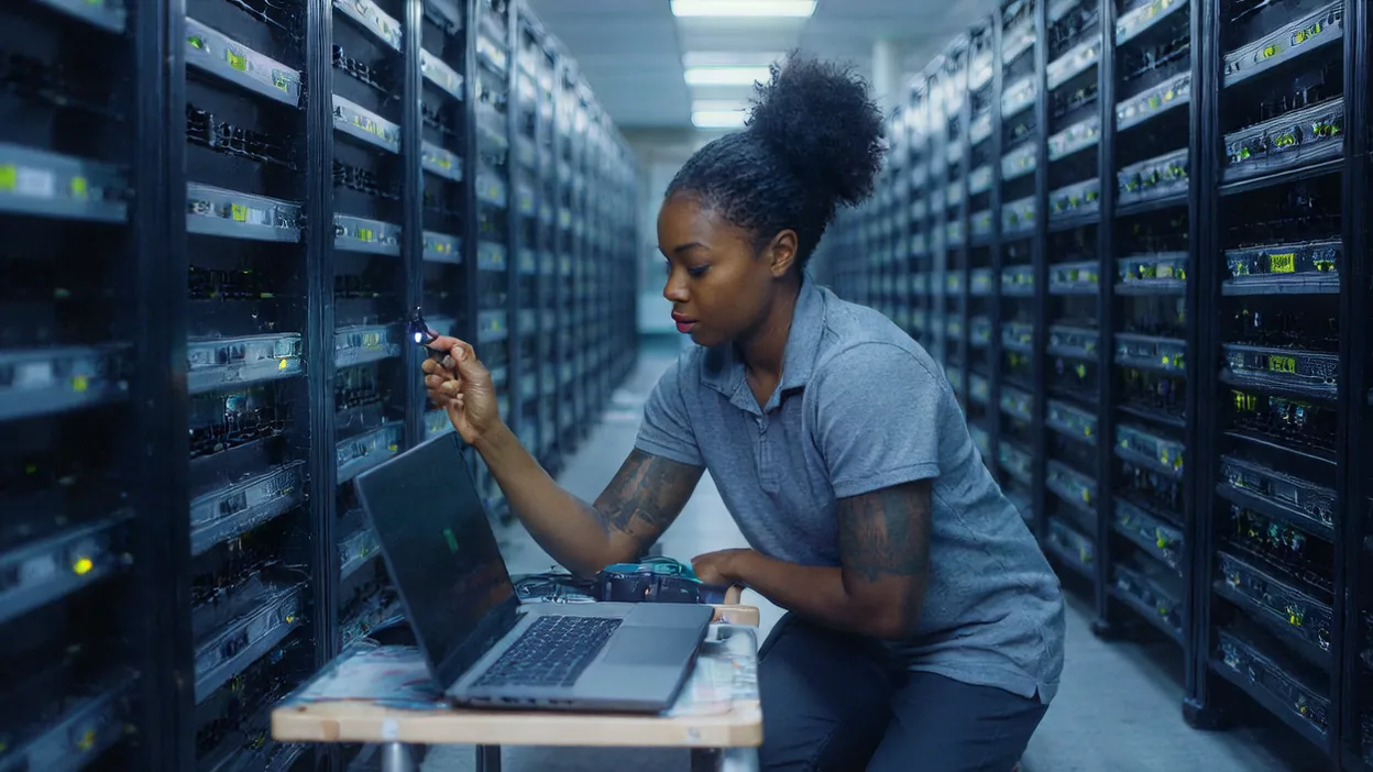 A technician uses a flashlight to inspect equipment in a long aisle of server racks, a laptop on a cart beside her.