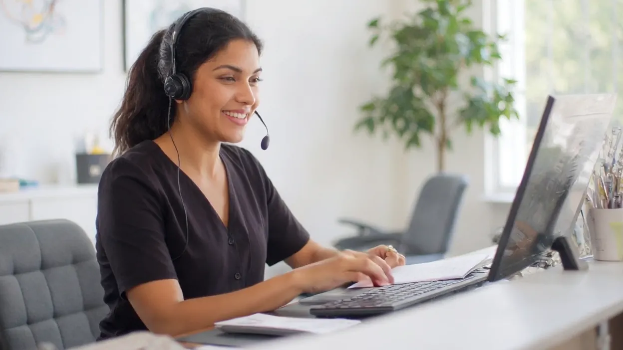 A smiling receptionist in a headset talks with a caller at her desk.