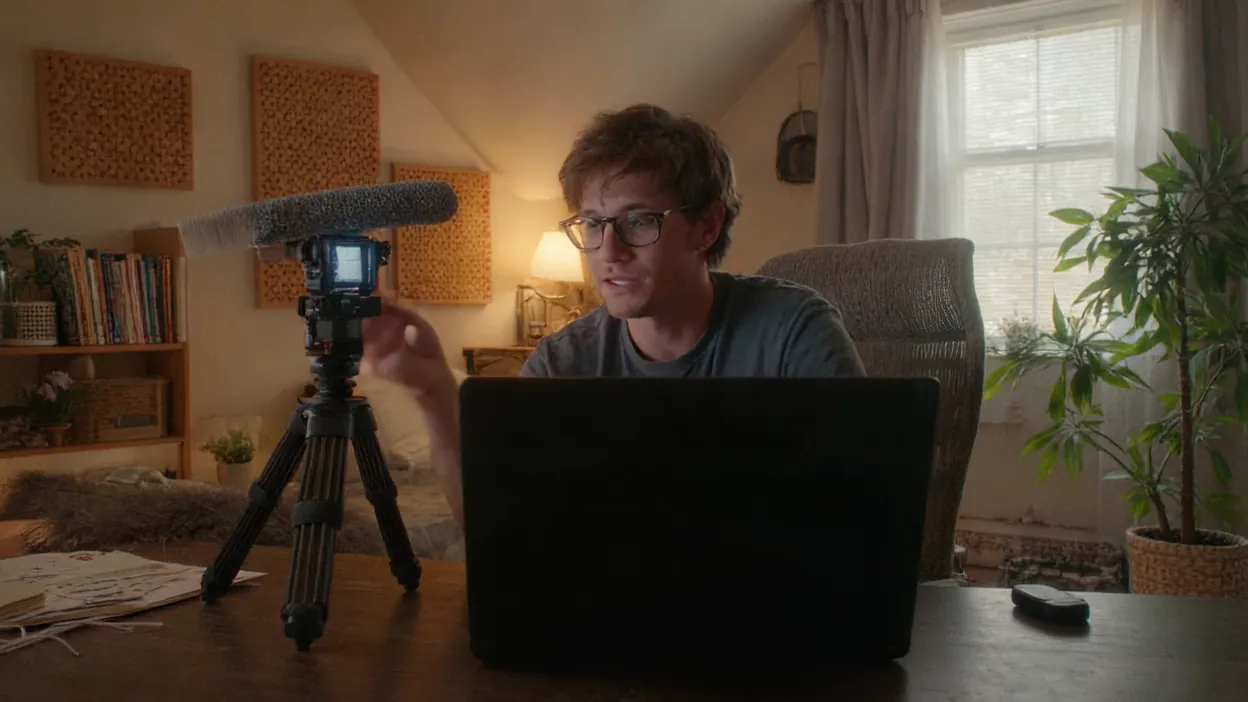 A freelance designer records a video with a camera on a tripod at his home office desk.