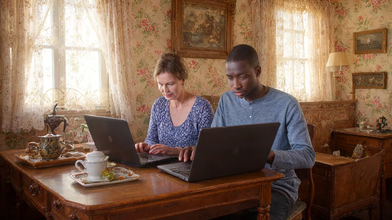 An inn owner and a web technician work side by side on laptops in a sitting room with floral wallpaper.