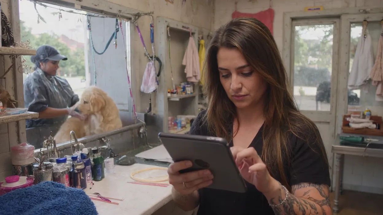 A dog groomer checks bookings on a tablet while a coworker bathes a golden retriever behind her.