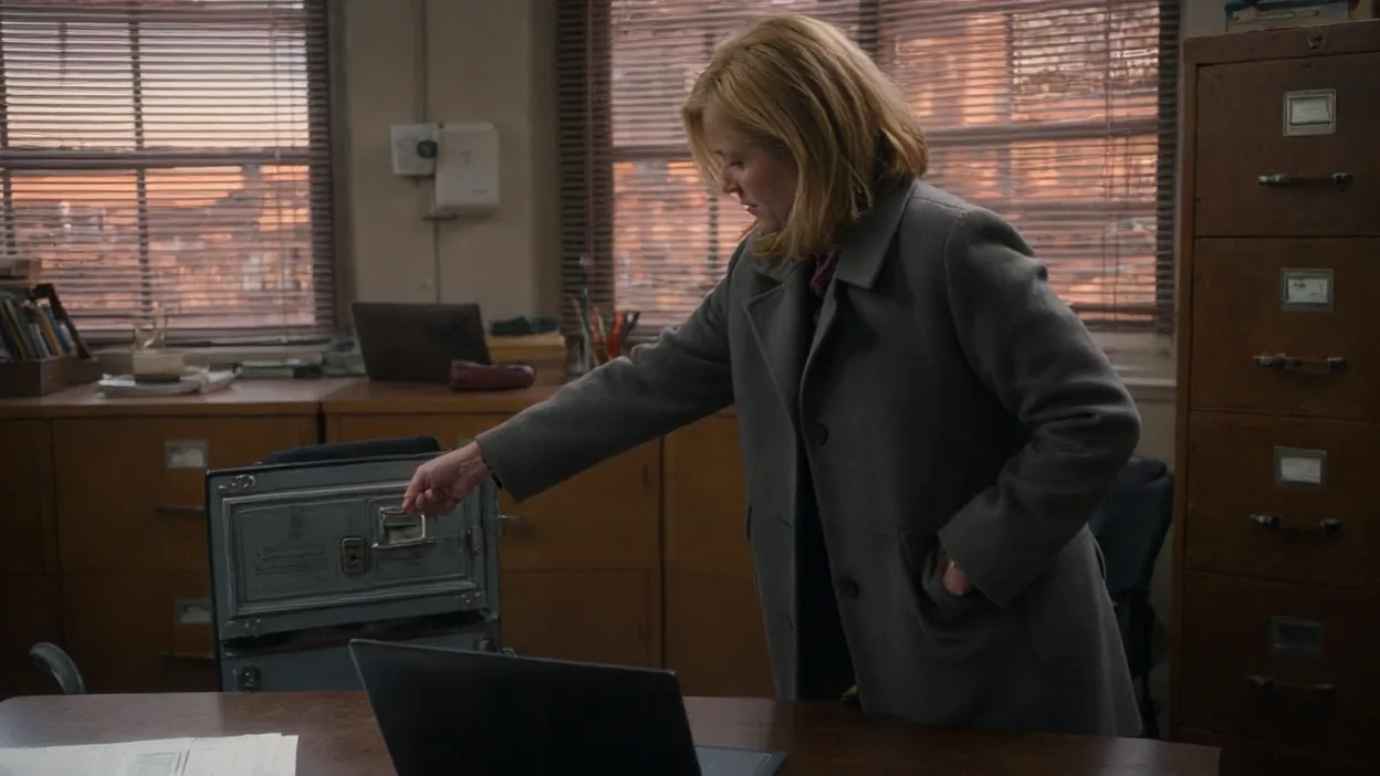A woman in a gray coat reaches for the handle of a metal safe beside her office desk at dusk, wooden filing cabinets and window blinds behind her.
