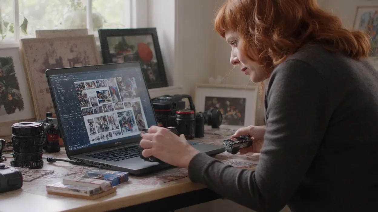 A photographer scrolls through a large folder of photos on a laptop at her desk among framed prints and cameras.