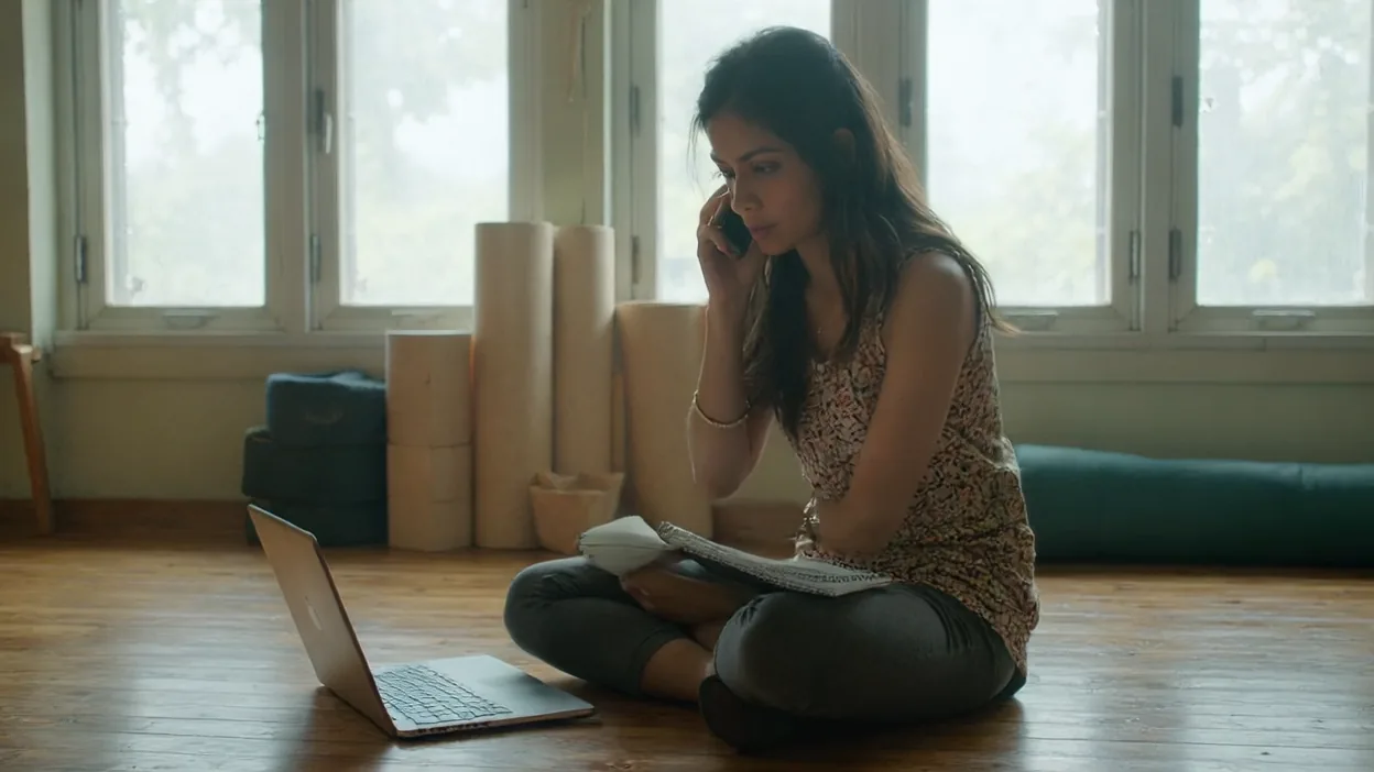 A yoga studio owner sits on the floor of her studio on a phone call, notes in her lap and a laptop beside her.